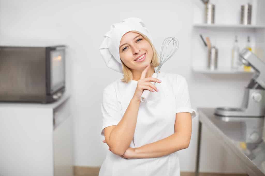 About Me 1 Smiling female chef in a white uniform and hat holding a whisk in a professional kitchen