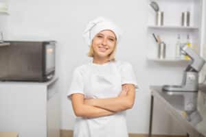 Confident female chef in a white uniform and hat, standing with arms crossed in a modern kitchen