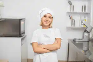 Confident female chef in a white uniform and hat, standing with arms crossed in a modern kitchen