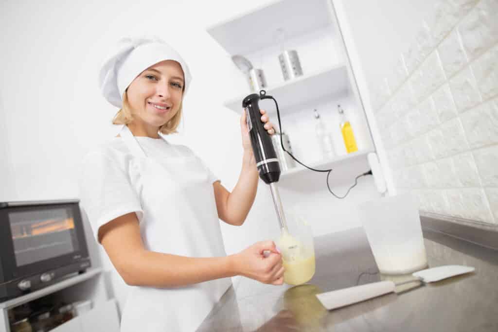About Me 2 Smiling female chef using a hand blender to mix ingredients in a professional kitchen