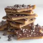 Protein Cookie Dough Bark That Hits Every Mark 10 Stack of chocolate-covered protein cookie dough bark pieces topped with sea salt and chocolate chips on a white surface.