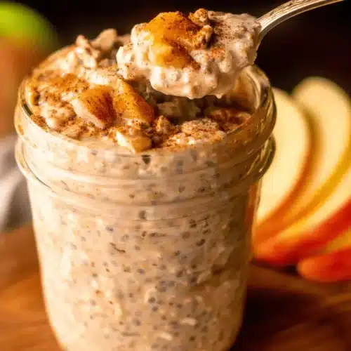 Protein Apple Cinnamon Overnight Oats (No Cook, High Fiber) 3 A spoon scoops creamy Apple Cinnamon Protein overnight oats from a glass jar, garnished with cooked apples and cinnamon, on a wooden board.