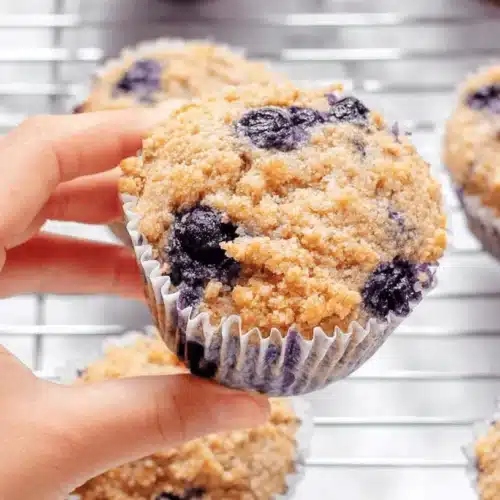 Close-up of a hand holding a freshly baked blueberry protein muffins with a golden streusel topping, alongside other muffins on a cooling rack.