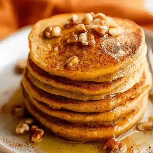 Close-up of a stack of golden-brown Pumpkin Protein Pancakes topped with walnuts and drizzled with glistening syrup.
