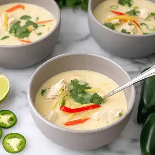 Three bowls of vibrant Thai chicken soup, garnished with fresh cilantro, lime, and peppers on a marble countertop.
