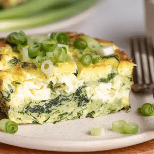 A close-up of a vibrant slice of baked cottage cheese eggs with spinach and fresh scallions on a rustic ceramic plate.