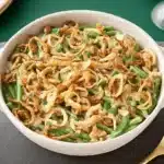 Rustic oval dish of homemade Green Bean Casserole with a crispy fried onion topping on an emerald green table.