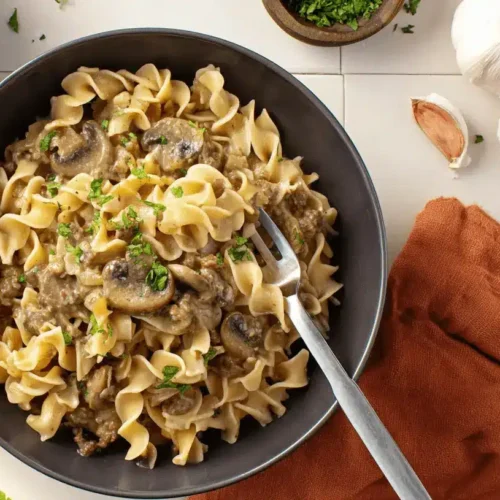 High-Protein Beef Stroganoff: A Healthier Twist on a Classic 3 Overhead view of a hearty High-Protein Beef Stroganoff with wavy egg noodles, mushrooms, and fresh parsley, served in dark bowls.
