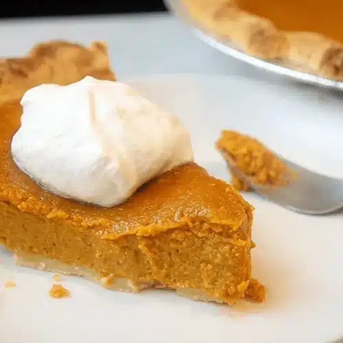 A delicious slice of southern sweet potato pie topped with whipped cream on a white plate, with a fork.