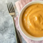 A warm bowl of vibrant, creamy orange sweet potato pudding with a fork on a marble surface.