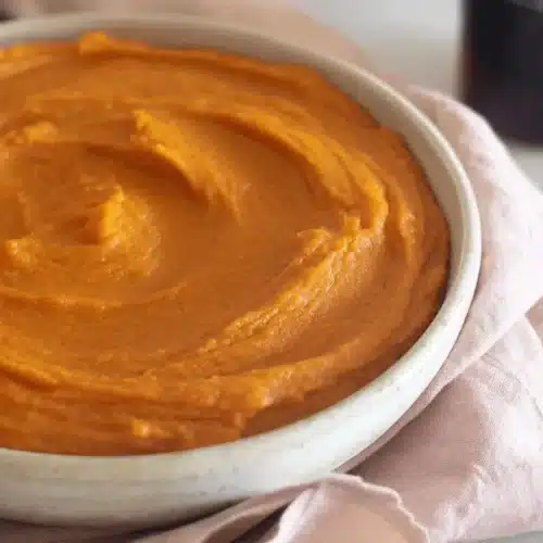 A warm bowl of vibrant, creamy orange sweet potato pudding with a fork on a marble surface and pink fabric.