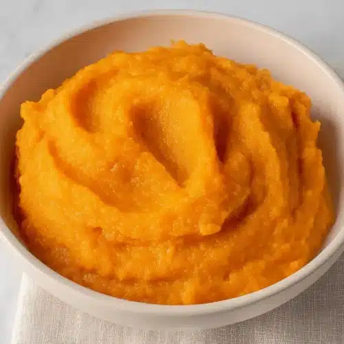 Velvety Brown Butter Sweet Potato Puree Recipe 2 A close-up, overhead view of a vibrant orange sweet potato puree in a light bowl on a marble and fabric surface.