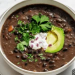 Beautifully garnished bowl of rich black bean soup with avocado, sour cream, and fresh cilantro, presented on a light surface.
