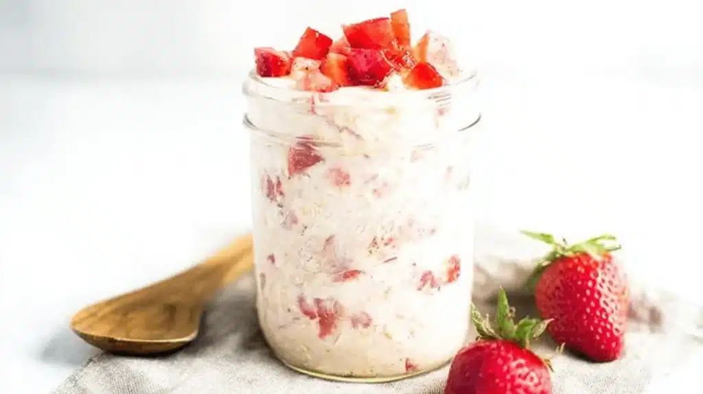 A bright still life of strawberry overnight oats in a mason jar, topped with fresh strawberries and accompanied by a wooden spoon and whole strawberries.