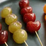 Vibrant candied fruit skewers, a delicious tanghulu recipe featuring grapes, strawberries, and mandarins on a dark platter.