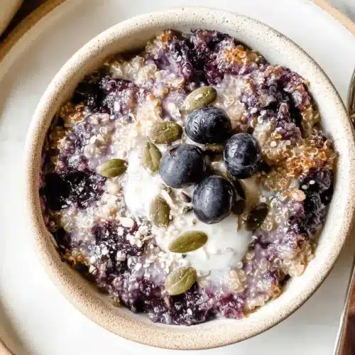 Delicious Blueberry Quinoa Breakfast Bake (Vegan & Gluten-Free) 2 A rustic bowl of homemade Blueberry Quinoa Breakfast Bake topped with fresh blueberries, creamy dollop, and pumpkin seeds on a white marble surface.