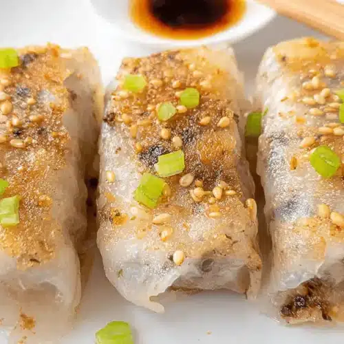 Close-up of three crispy pan-fried rice paper dumplings garnished with sesame seeds and green onions on a white plate with dipping sauce.