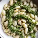 Close-up of creamy pesto beans garnished with red chili flakes and fresh basil in a distressed metallic bowl.