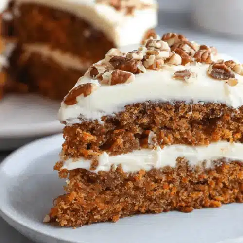 Easy & Moist Carrot Cake with Cream Cheese Frosting 2 A delicious slice of multi-layered carrot cake with cream cheese frosting and chopped pecans on a plate, with a whole carrot cake in the blurred background.