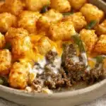 Hearty tater tot casserole with ground beef, golden tots, green beans, and melted cheese in a rustic baking dish.