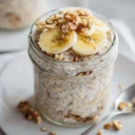 Banana bread overnight oats topped with banana slices, chopped walnuts, and honey in a glass jar.