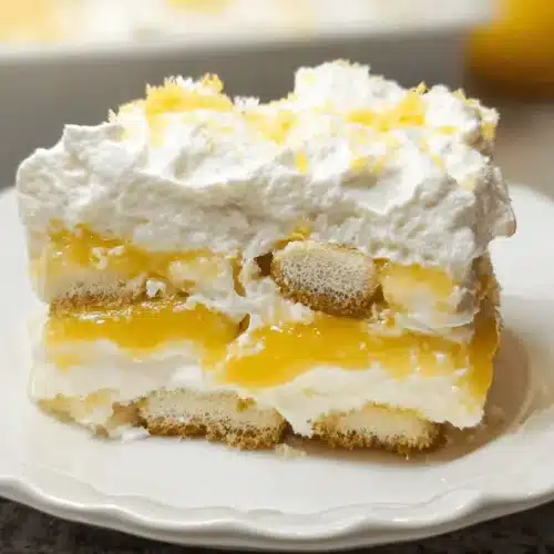 A delicious slice of layered lemon tiramisu recipe, with creamy white layers, vibrant yellow lemon curd, and ladyfingers, garnished with lemon zest on a white scalloped plate.