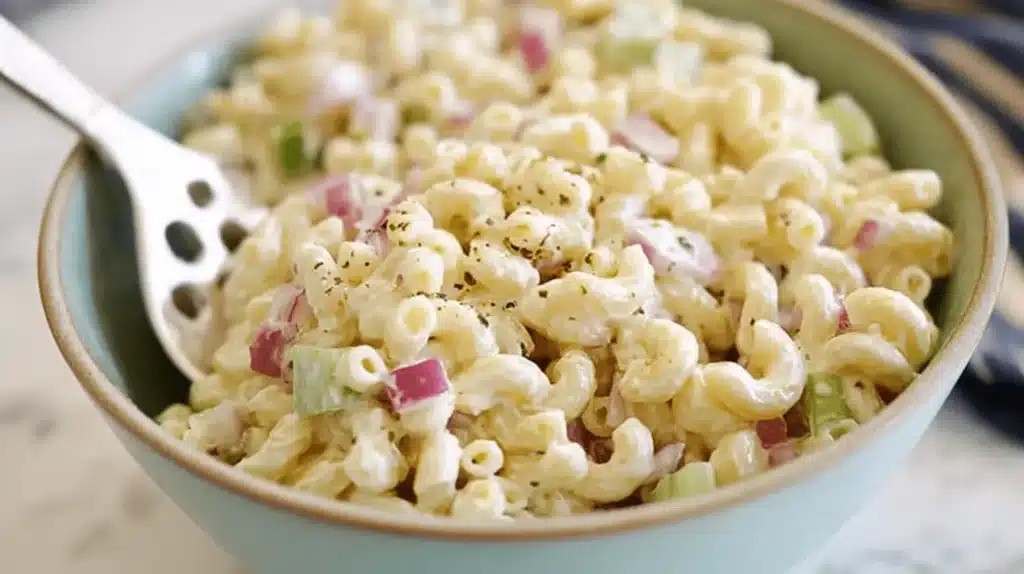 Creamy macaroni salad recipe served in a rustic bowl with red onions, celery, and black pepper.