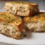 Three golden-brown grilled tuna melt sandwiches with gooey cheese and fresh herbs on a white plate, showcasing a delicious tuna melt recipe.