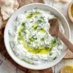 Overhead view of a creamy homemade tzatziki sauce recipe garnished with dill and olive oil, served with pita bread and lemon on a rustic wooden table.